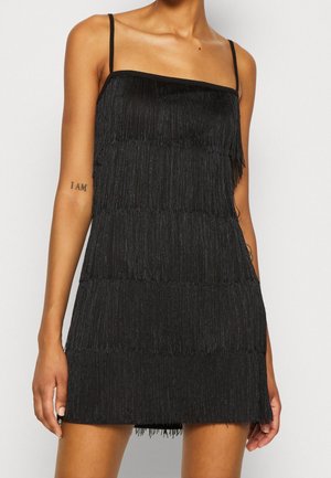 Vrouw draagt een zwarte mouwloze jurk met franjes en dunne bandjes, toont tatoeages op beide bovenarmen en staat tegen een effen achtergrond.