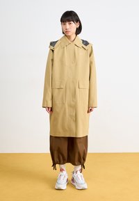 Manteau long beige clair avec un col, deux poches avant et des accents texturés aux épaules. Le modèle porte un pantalon marron et des baskets.