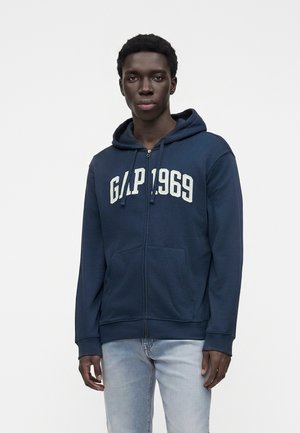 Joven con sudadera con capucha azul marino con cremallera y "GAP 1969" en letras blancas, y jeans azul claro, de pie contra un fondo blanco liso.
