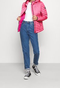 Ljusrosa quiltad jacka med huva, svart dragkedja och logotypmärke, kombinerad med högmidjade blå jeans och svarta sneakers.