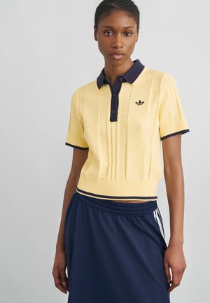 Mujer joven que lleva una camiseta de polo de punto amarilla de manga corta con cuello azul marino y falda azul marino con cintura elástica.