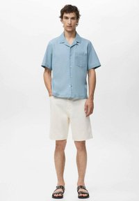 Camisa de boton corto de manga corta en azul claro con un bolsillo, combinada con pantalones cortos color crema y sandalias negras. Diseño simple y tejido ligero.