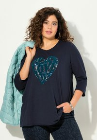 Marineblaues Langarmshirt mit einem großen grafischen Teal-Herz; kombiniert mit einer hellen Teal-Steppjacke und gemusterten dunklen Leggings.