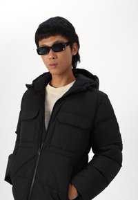 Svart jakke med hette, med to brystlommer og glidelås. Modellen har på seg svarte solbriller og en beige topp under.
