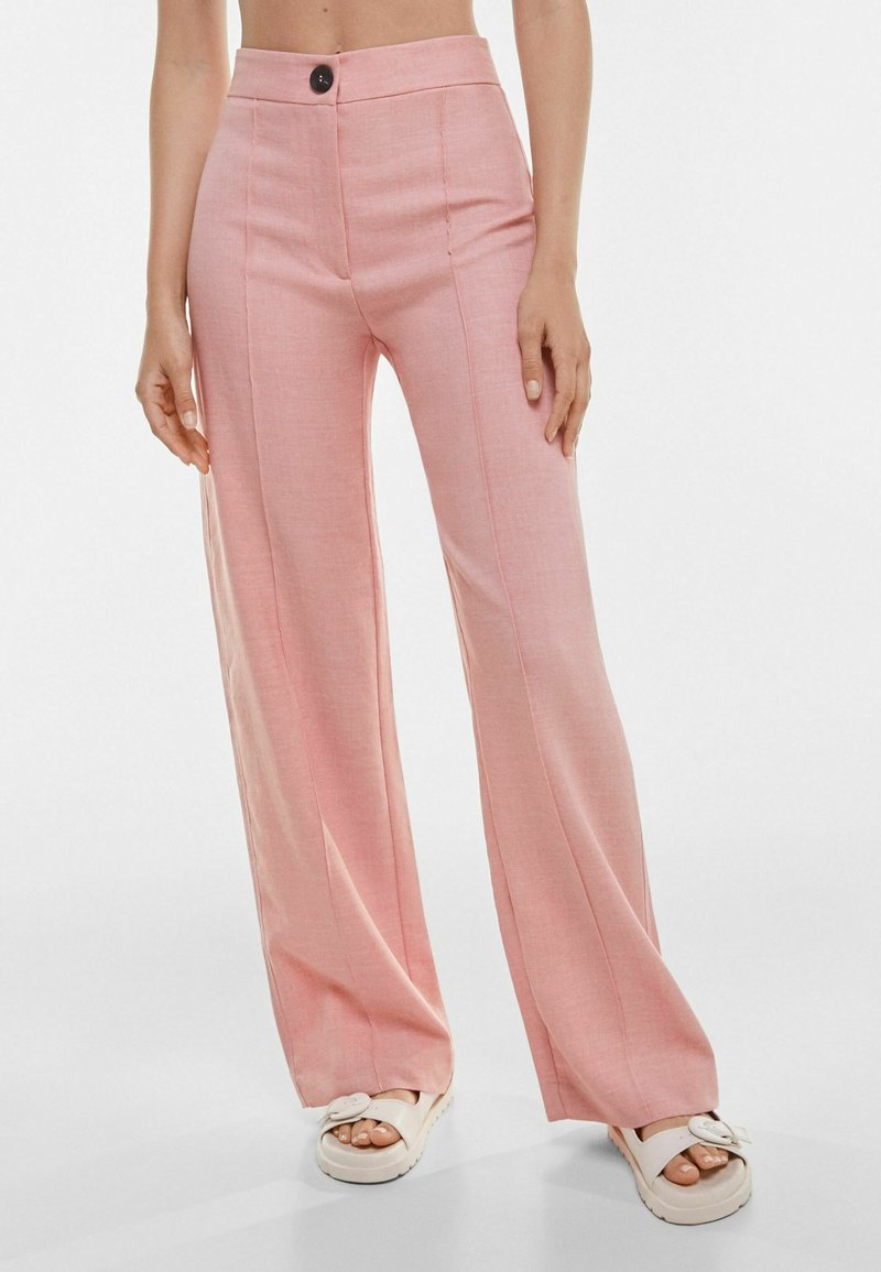 Personne portant un pantalon rose taille haute à jambes larges avec un bouton noir et des sandales blanches sur fond blanc.