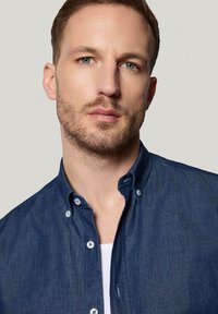 Ciemnoniebieska koszula dżinsowa z kołnierzykiem na guziki, z jasnymi guzikami i miękką teksturą, noszona na białej koszulce.