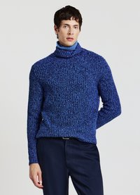 Blauer Rollkragenpullover mit strukturierter, grobgestrickter Optik. Lange Ärmel und lockerer Schnitt. Kombiniert mit dunklen Hosen, die einen Lagenlook zeigen.