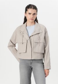 Veste beige courte en tissu léger, avec un grand col, deux poches avant et des cordons au niveau du cou.