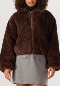 Bruine, fuzzy, cropped jas met een rits aan de voorkant, brede mouwen en een gekraagde halslijn, gecombineerd met een grijze rok met twee voorzakken.