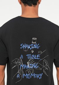 Persona, tērpusies melnā kreklā ar zilo tekstu "Kopā pie galda, veidojot atmiņu" un baltām līniju zīmējumiem ar rokām, kas tur kartes.