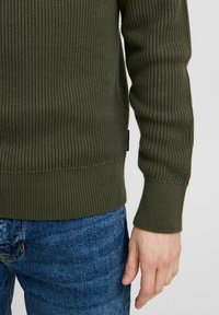 Grüner Rippstrickpullover mit strukturierter Oberfläche. Verfügt über lange Ärmel und einen körpernahen Saum. Kombiniert mit blauen Denim-Jeans.