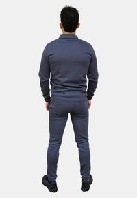 Tuta grigio scuro realizzata in un morbido tessuto testurizzato; presenta un colletto con zip e un design aderente. Abbinato a sneaker nere. Vista posteriore.