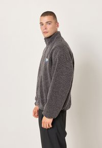 Jaunas vyras, dėvintis pilką užtraukiamą fliso striukę su mažu spalvotu logotipu ant krūtinės, stovintis profilio pusėje priešais vientisą foną.