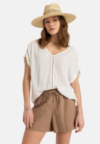 Blusa bianca testurizzata con scollatura a V e dettagli sulla chiusura, abbinata a pantaloni corti marroni con cordoncino realizzati in un tessuto leggero. Cappello di paglia che completa il look.