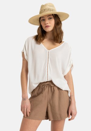 Blusa bianca testurizzata con scollatura a V e dettagli sulla chiusura, abbinata a pantaloni corti marroni con cordoncino realizzati in un tessuto leggero. Cappello di paglia che completa il look.