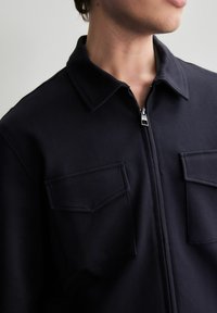 Chaqueta azul marino de cierre tipo cremallera, fabricada en tejido texturizado, con un cuello y dos bolsillos frontales con solapas. Los detalles incluyen una cremallera plateada.