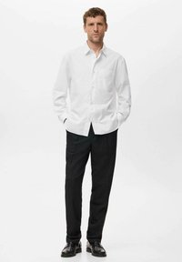 Camisa de botones blanca a rayas con un bolso en el pecho, combinada con pantalones de vestir negros y zapatos negros. El modelo está de pie con las manos en los bolsillos.