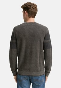 Man in een donkergrijze gebreide trui met lange mouwen en donkerdere elleboogpatches, gecombineerd met blauwe jeans, met de rug naar de camera gekeerd tegen een witte achtergrond.