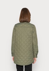 Persona de espaldas vistiendo una larga chaqueta acolchada verde oliva con costuras en forma de diamante y pantalones negros, frente a un fondo simple.