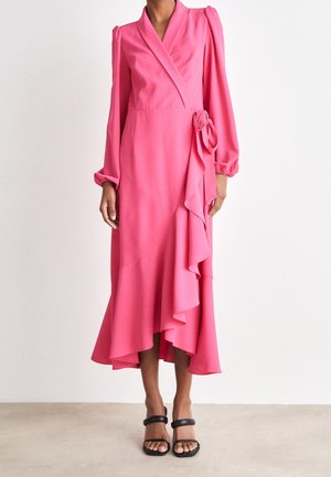 Femme portant une robe portefeuille rose à manches longues avec un volant asymétrique et des sandales à talons noires, debout devant un fond uni.