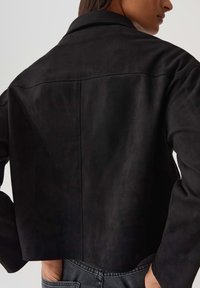 Giacca nera cropped in tessuto morbido con colletto classico, dettagli di cucitura sul retro e maniche lunghe, abbinata a jeans di denim.