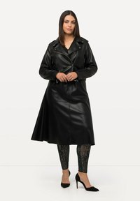 Schwarzer Leder-Trenchcoat mit tailliertem Gürtel, ausgestattet mit einem Reverskragen und Reißverschlusstaschen, kombiniert mit gemusterten schwarzen Leggings und High Heels.