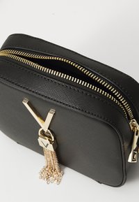 Svart strukturerad crossbody-väska med guld blixtlås, V-formad guld hårdvara och hängande guld kedjetasslar mot en vit bakgrund.