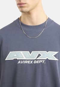 Szara bawełniana koszulka z dużym, haftowanym niebiesko-białym logo z napisem "AVIREX DEPT.", okrągłym dekoltem i teksturowanym materiałem.