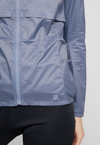 Coupe-vent léger bleu avec une fermeture éclair à l'avant, deux poches poitrine et un logo Salomon en bas à gauche. Texture lisse et ripstop.