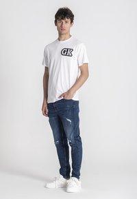 Biała koszulka z krótkim rękawem z dekoracyjnym czarnym logo "GK", w połączeniu z postarzanymi niebieskimi dżinsami i białymi sneakersami.