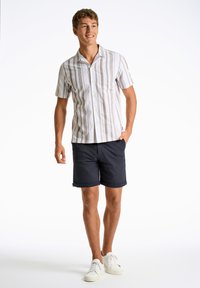 Camisa de rayas de manga corta en blanco y marrón claro, combinada con pantalones cortos azul marino. Modelo usando zapatillas blancas, de pie sobre un fondo liso.