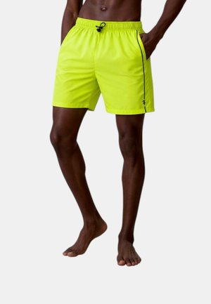Uomo dalla pelle scura che indossa pantaloncini giallo brillante con coulisse nera e striscia laterale, in piedi a piedi nudi su uno sfondo semplice.