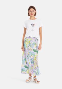 Weißes T-Shirt mit Cocktailgrafik, kombiniert mit einem fließenden, mehrfarbigen Maxirock mit lebhaften Drucken und Mustern, getragen mit weißen Sandalen.