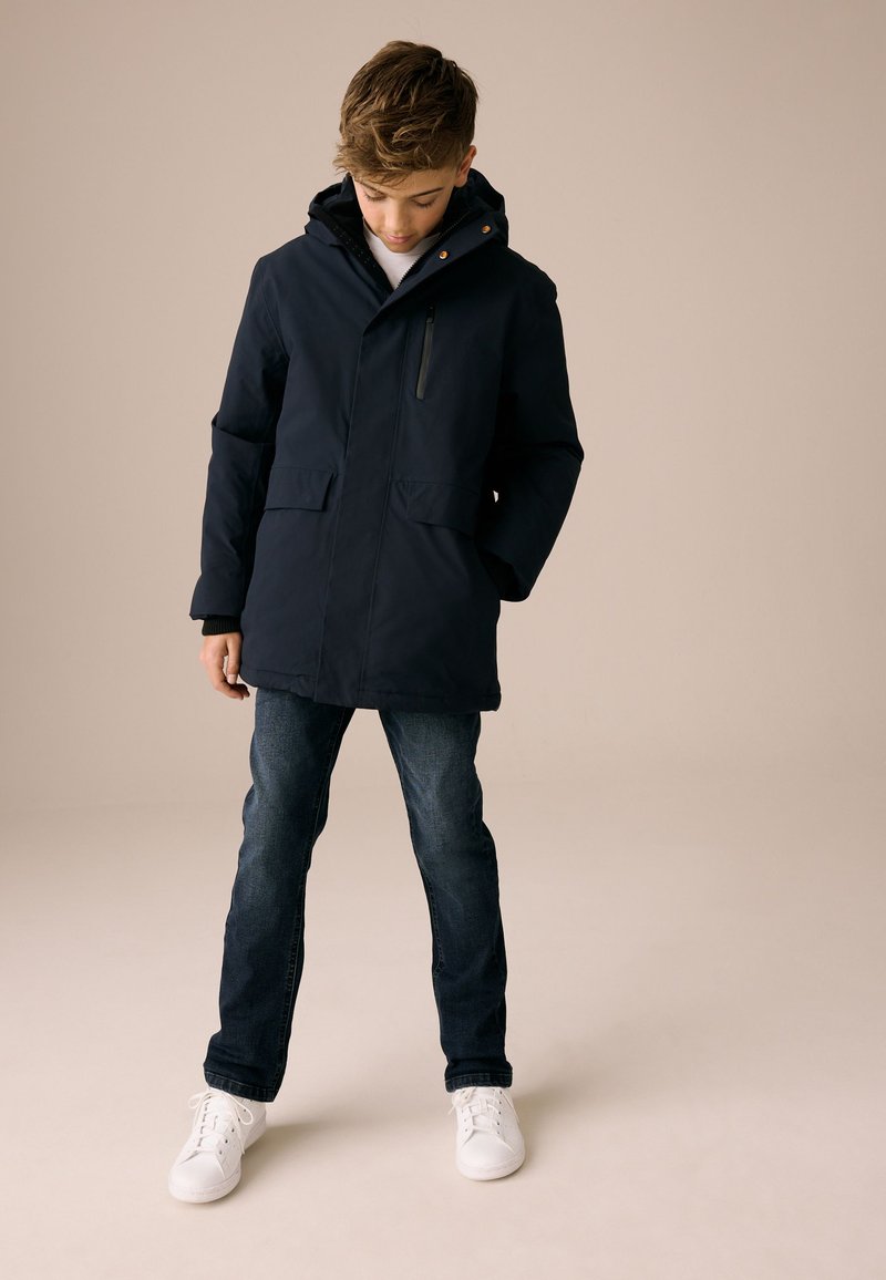 Giacca imbottita blu navy con cappuccio, tasche frontali e tasca con zip. Abbinata a jeans scuri e scarpe da ginnastica bianche. Texture liscia.