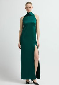 Dlouhé zelené saténové šaty bez rukávů s vysokým límcem a rozparkem na stehně, s hladkou strukturou a přiléhavým střihem, doplněné černými podpatky.