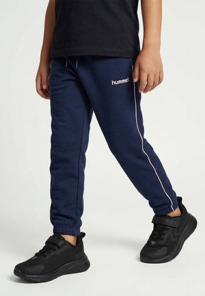 Enfant portant un pantalon de survêtement hummel bleu marine avec des bandes roses et des baskets noires, debout sur un fond blanc.