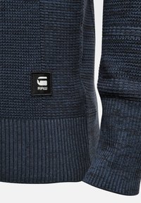 Manche et ourlet d’un pull en maille texturée bleu foncé avec une étiquette rectangulaire noire affichant un logo blanc « G RAW ».