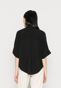 Blusa nera a maniche corte con una vestibilità morbida, orlo arrotondato e dettagli plissettati sulla schiena. Realizzata in un tessuto leggero con una texture liscia.