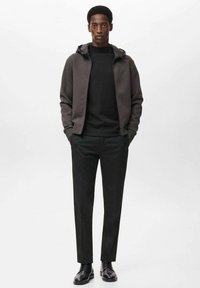 Hanorac gri cu fermoar, cu interior negru, purtat deasupra unui pulover negru, asortat cu pantaloni negri eleganți și pantofi negri.