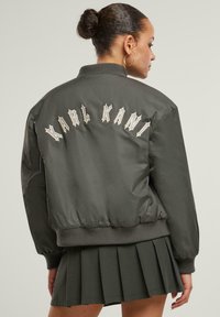 Khaki-Bomberjacke mit gerippten Bündchen, auf deren Rückseite "KARL KANI" in Creme gestickt ist. Hergestellt aus schimmerndem, glattem Material.