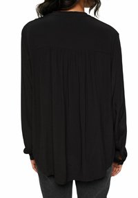 Blouse noire à manches longues en tissu doux, avec un dos plissé et un col rond. Agrémentée de détails au niveau des poignets.