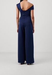 Mono azul marino con diseño off-shoulder, cuerpo ajustado, piernas anchas y cierre de cremallera en la parte posterior. Textura de tela suave.