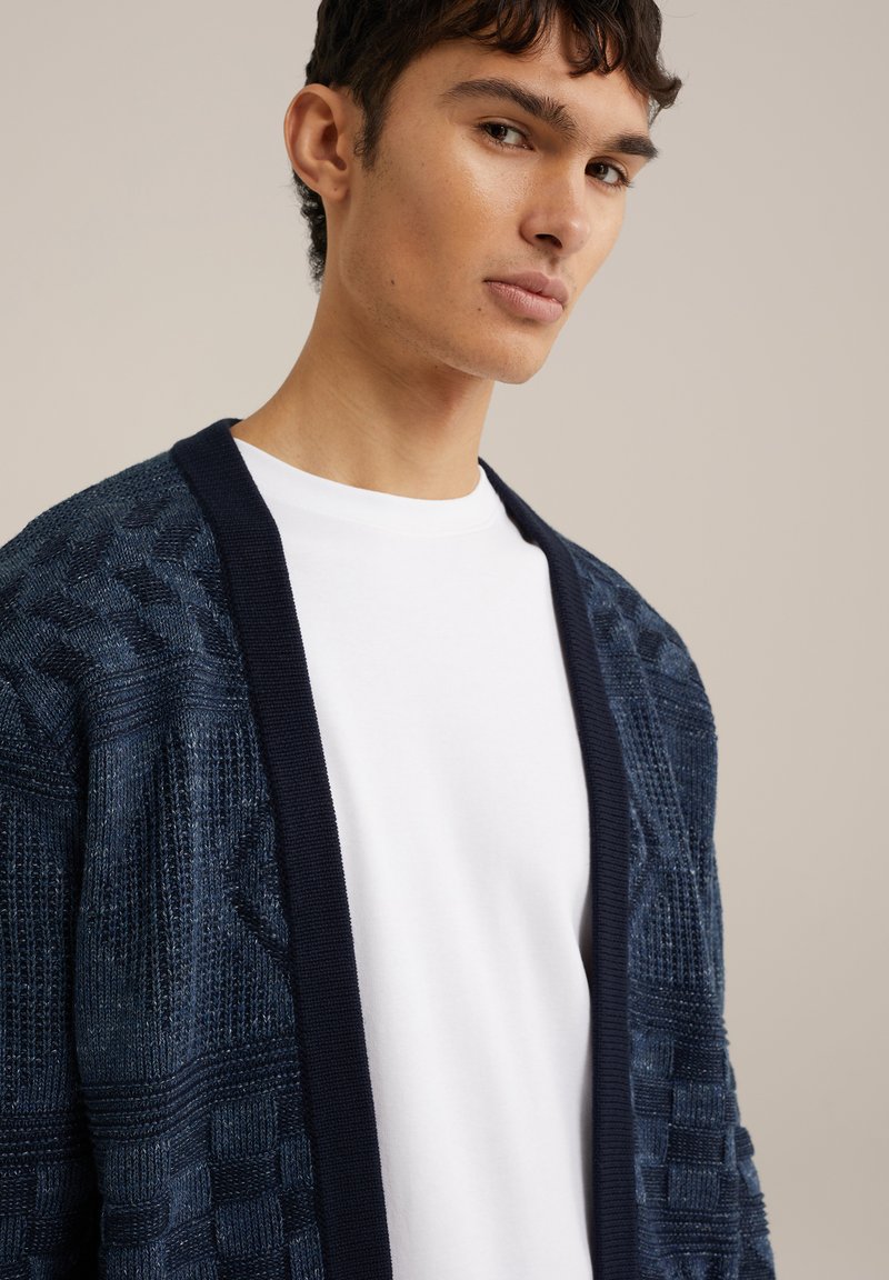 Navyblauwe textuur cardigan met een gebreid patroon en contrasterende donkerblauwe rand, laag over een wit T-shirt.