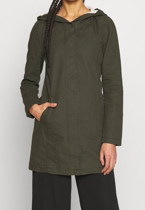 Mujer con abrigo con capucha de color verde oscuro con bolsillos, con la mano izquierda dentro de un bolsillo y el brazo derecho relajado a un lado.