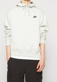 Ljusgrå hoodie med dragsko i huvan, framficka, ribbade muddar och en liten svart Nike-logga på bröstet; mjukt material, avslappnad passform.