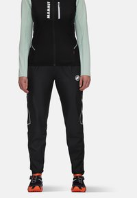 Svarta träningsbyxor med reflexdetaljer, zipfickor, kombinerat med en svart väst och långärmad mintgrön tröja, bärs tillsammans med svarta och orange skor.