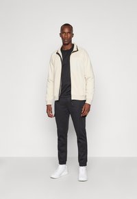 Giacca zip beige chiaro con tasca sul petto, abbinata a pantaloni jogger neri e sneakers bianche. Tessuto morbido e vestibilità rilassata.