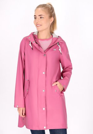 Vrouw draagt een roze regenjas met capuchon over een roze-wit gestreept shirt, met één hand in de zak, glimlachend en zijwaarts kijkend.