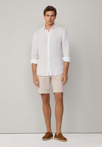 White linen button-up shirt, beige shorts, and brown slip-on shoes. The shirt has rolled sleeves and a small emblem on the chest.
