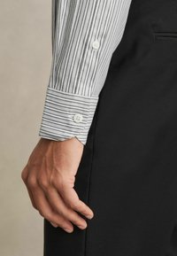 Cămașă cu mânecă lungă, cu dungi gri și albe, având o manșetă cu buton. Țesătura pare netedă, asortată cu pantaloni negri.